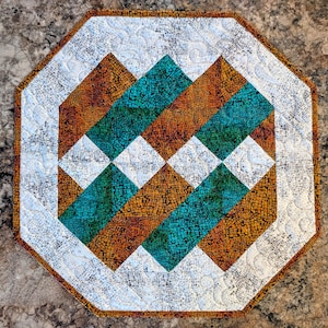 May include: An octagonal quilted table topper with a geometric design. The pattern features alternating diagonal rows of teal and orange textured fabric, separated by white diamond shapes. The border is white with a subtle pattern.