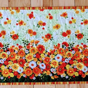 May include: A rectangular floral table runner featuring a vibrant design. The runner showcases a gradient of colorful flowers, transitioning from orange, yellow, and white at the bottom to a lighter green background with scattered flowers. The edges are bordered with a striped pattern.