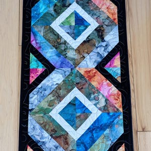May include: A colorful table runner with a geometric design featuring a black border and white diamond shapes. The runner is made of fabric with a batik-style pattern in shades of blue, green, orange, pink, and brown.