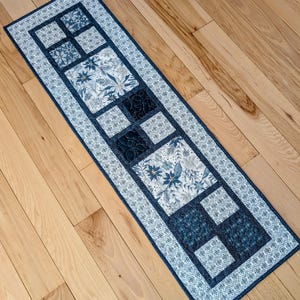 May include: A rectangular quilted table runner with a repeating pattern of blue and white squares. The squares are bordered by a patterned blue and white fabric. The center squares feature floral and geometric designs. The runner is placed on a wooden surface.