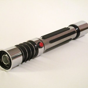 May include: A silver and black lightsaber prop with a red button. The lightsaber has a textured black grip and a silver metal end cap.