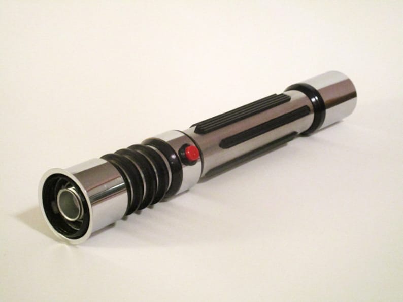 May include: A silver and black lightsaber prop with a red button. The lightsaber has a textured black grip and a silver metal end cap.