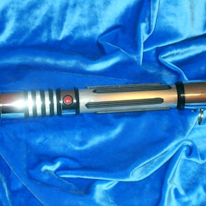 May include: A silver and black lightsaber with a red button on the handle. The lightsaber is on a blue velvet background.