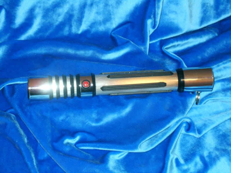 May include: A silver and black lightsaber with a red button on the handle. The lightsaber is on a blue velvet background.
