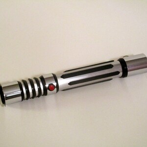 May include: A silver and black lightsaber with a red button. The lightsaber has a textured grip and a silver blade.