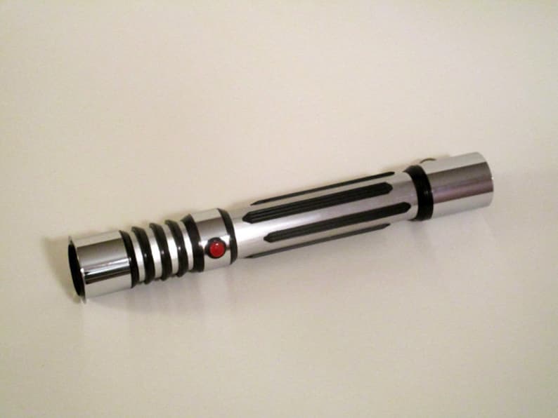 May include: A silver and black lightsaber with a red button. The lightsaber has a textured grip and a silver blade.