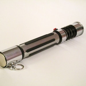 May include: A silver and black lightsaber keychain with a red button. The keychain has a silver cap on one end and a black and silver handle with a black spring.