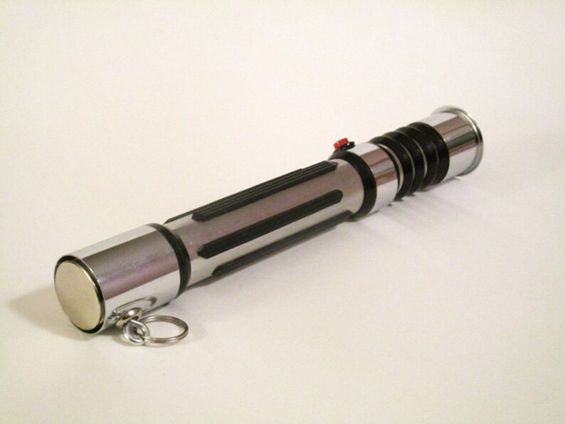 May include: A silver and black lightsaber keychain with a red button. The keychain has a silver cap on one end and a black and silver handle with a black spring.
