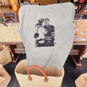 May include: A light gray t-shirt featuring a black and white image of two hockey players embracing. The shirt is displayed over a woven basket with a brown leather handle. The background includes other baskets and store displays.