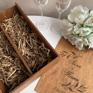 Könnte beinhalten: Zwei Sektgläser mit goldenen Kronen und dem Wort "Mrs" werden gezeigt. Eine Holzkiste mit geschreddertem Papier und ein Holzschild mit Namen und Datum sind ebenfalls vorhanden. Ein Strauß weißer Blumen befindet sich neben den Gläsern.