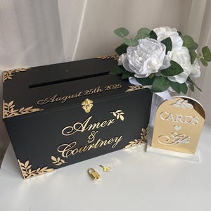May include: Black wooden wedding card box with gold accents and a lock and key. The box has the names "Amer & Courtney" and the date "August 25th 2025" engraved on it. A gold mirror sign reads "Cards & Gifts".