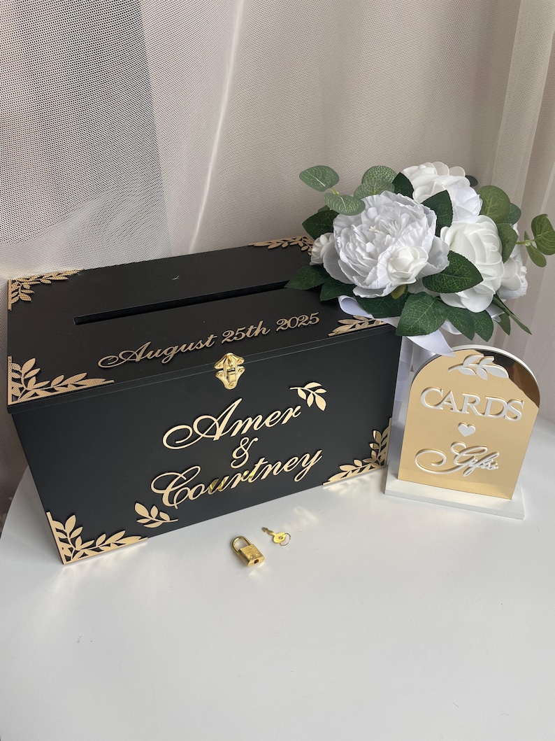 May include: Black wooden wedding card box with gold accents and a lock and key. The box has the names "Amer & Courtney" and the date "August 25th 2025" engraved on it. A gold mirror sign reads "Cards & Gifts".