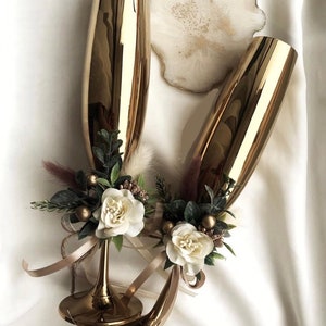 Peut inclure: Deux flûtes à champagne dorées ornées de fleurs blanches et de verdure attachées autour des tiges. Les flûtes sont sur une surface blanche.