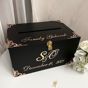 May include: Black wooden box with a gold lock and key. The box has a slot on the top for cards. The box is decorated with gold filigree and the text "Family Gruberuk, S&O, December 19, 2023".