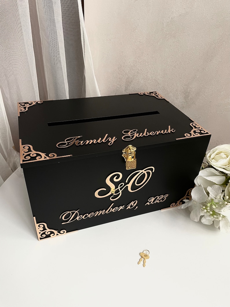 May include: Black wooden box with a gold lock and key. The box has a slot on the top for cards. The box is decorated with gold filigree and the text "Family Gruberuk, S&O, December 19, 2023".