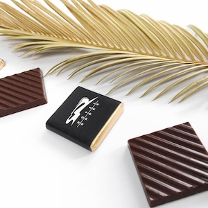 May include: Three chocolate bars, two are plain dark chocolate with a ribbed texture, and one is wrapped in black foil with a gold trim and a white logo that says "HOTEL" with four stars.