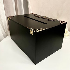 May include: Black wooden box with a keyhole and ornate gold metal corners. The box has a slot on the top for inserting cards. The box is sitting on a white surface with a gold key next to it.