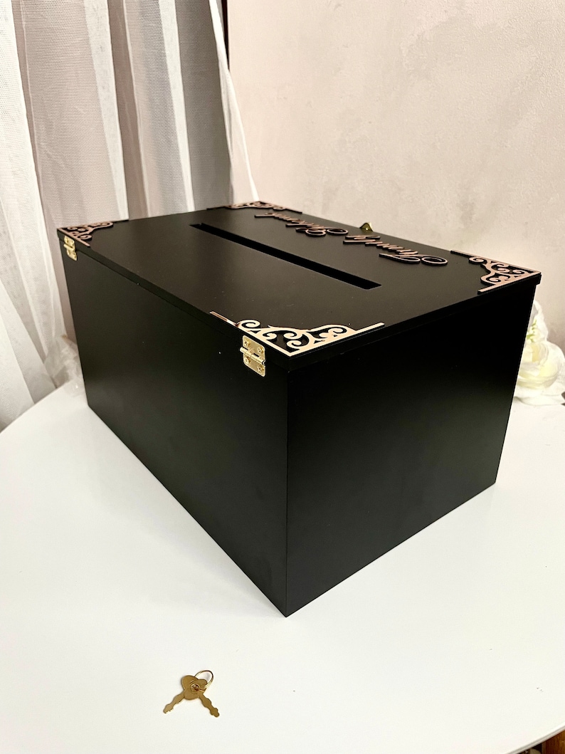 May include: Black wooden box with a keyhole and ornate gold metal corners. The box has a slot on the top for inserting cards. The box is sitting on a white surface with a gold key next to it.