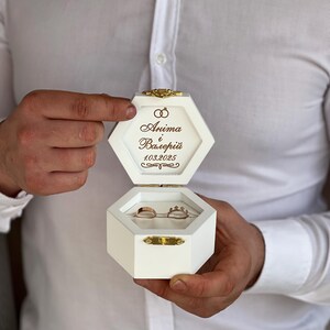 Könnte beinhalten: Weiße sechseckige Ringbox mit goldenem Verschluss und der Inschrift "Anima & Vasileii 10.3.2025". Zwei goldene Eheringe befinden sich in der Box.