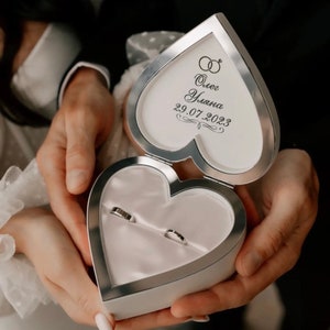 May include: A white heart-shaped ring box with a silver trim. The lid is open and has the inscription "Олег Ульяна 29.07.2023" on it. Two gold wedding bands are inside the box.