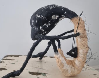May include: A sculptural figure with a black, textured head and limbs, hunched over a curved, beige fabric-wrapped body. The figure holds a small, black triangular pendant with a single silver-coloured bead, featuring loose dark threads.