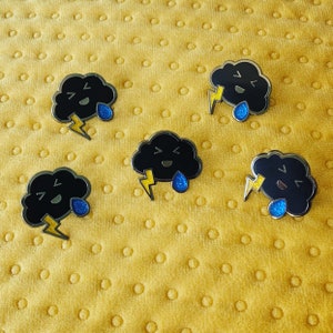 May include: Five black enamel pins shaped like clouds with a lightning bolt and a raindrop. Each cloud has a different facial expression.