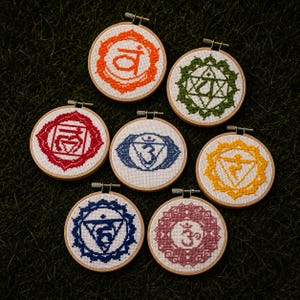 May include: Seven circular wooden embroidery hoops containing white fabric with cross-stitched chakra symbols in orange, green, red, blue, yellow, dark blue, and magenta. Each hoop features a metal tension screw at the top for display.