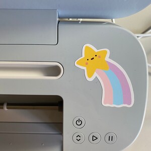 May include: A light blue cutting machine with a yellow star sticker with a rainbow tail. The machine has a power button, an arrow pointing up, an arrow pointing right, and a pause button.