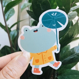 May include: A sticker of a cartoon frog wearing a yellow raincoat and holding a blue umbrella. The frog is smiling and has pink cheeks.