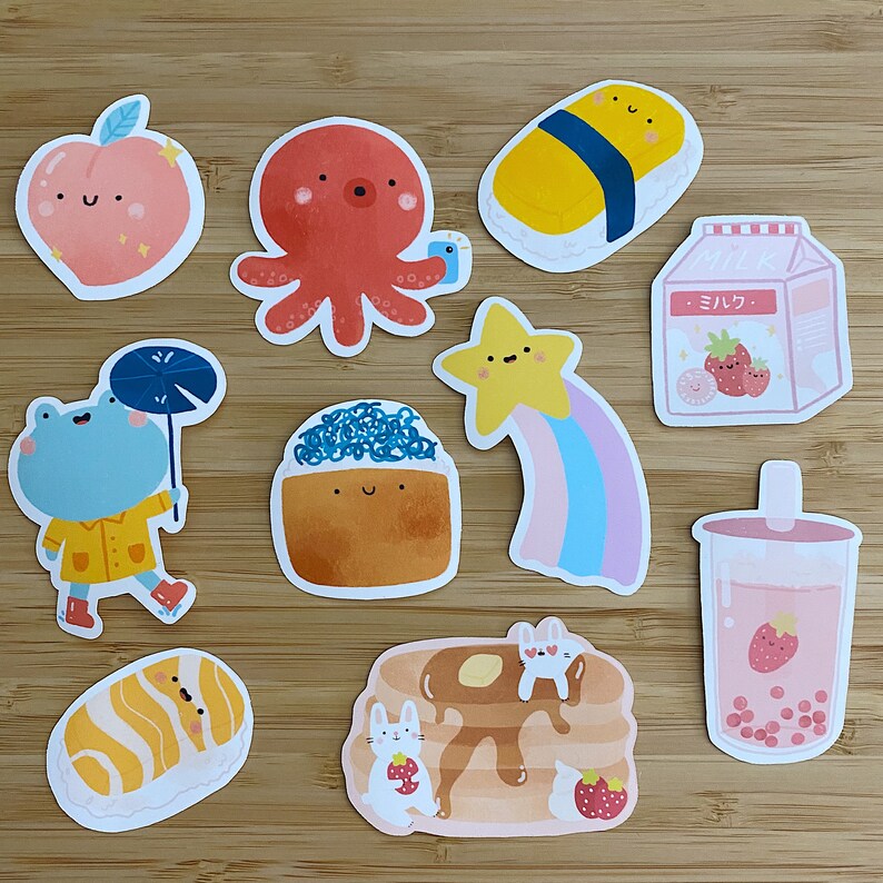 May include: A set of ten colorful stickers featuring various designs, including a red octopus, a yellow star with a rainbow tail, a pink apple with a smiling face, a blue frog wearing a raincoat, a sushi roll, a milk carton with the word "ミルク" (milk) in Japanese, a pancake with strawberries, a glass of boba tea, and a sushi roll.