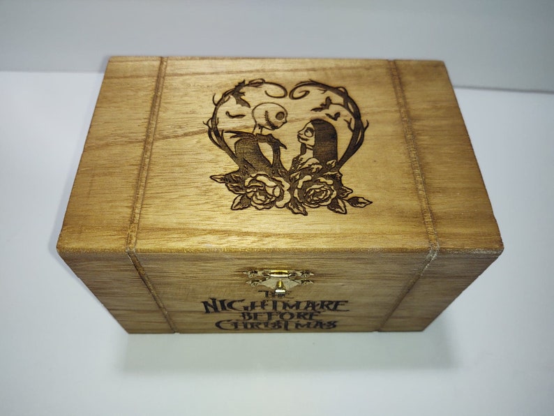 May include: A wooden box with a hinged lid. The lid is decorated with an engraved image of Jack Skellington and Sally from the movie "The Nightmare Before Christmas". The box is labeled "The Nightmare Before Christmas".