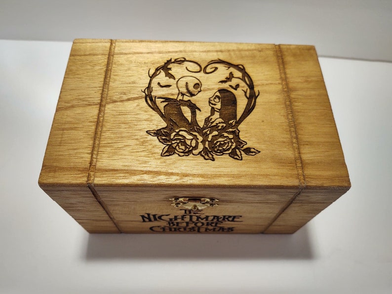 May include: A wooden box with a hinged lid. The top of the box is decorated with a laser-engraved image of Jack Skellington and Sally from the movie "The Nightmare Before Christmas". The text "The Nightmare Before Christmas" is engraved on the front of the box.