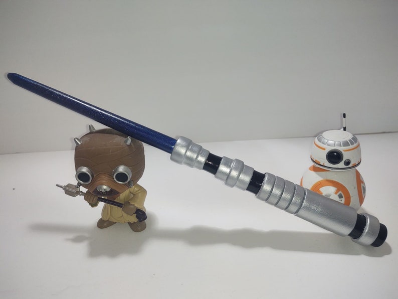 Op de afbeelding: Een blauw en zilveren lightsaber-speelgoed met een zwarte handgreep. De lightsaber ligt op een wit oppervlak naast een bruin en oranje speelgoedrobot.