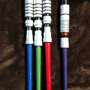 Op de afbeelding: Vier zilveren lightsaber-handvatten met zwarte accenten. De handvatten hebben blauwe, groene, rode en paarse bladen.