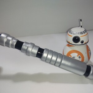 Op de afbeelding: Een zilveren en zwarte lightsaber-prop met een blauwe handgreep. De lightsaber ligt op een wit oppervlak naast een klein oranje en wit robot speelgoed.