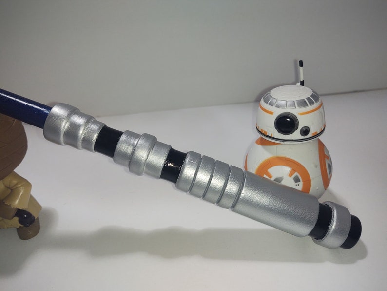 Op de afbeelding: Een zilveren en zwarte lightsaber-prop met een blauwe handgreep. De lightsaber ligt op een wit oppervlak naast een klein oranje en wit robot speelgoed.