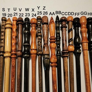 May include: A collection of 20 wooden wands with various wood grains and finishes. The wands are arranged in a row from left to right, with the tips of the wands pointing towards the top of the image. The wands are labeled with letters and numbers from Q to JJ.
