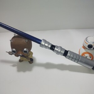 Op de afbeelding: Een zilveren en zwart lichtzwaard met een blauwe kling. Het lichtzwaard ligt op een wit oppervlak. Op de achtergrond zijn een klein bruin speelgoedfiguur en een witte en oranje speelgoedrobot te zien.