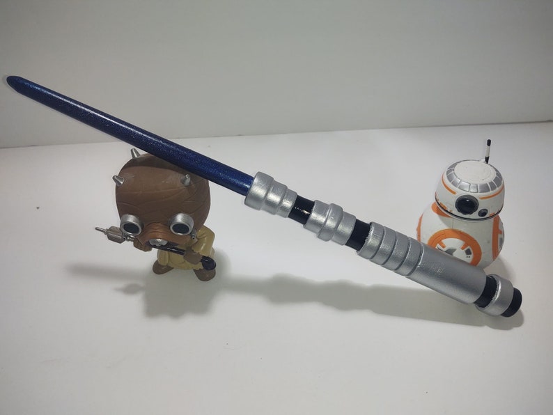 Op de afbeelding: Een zilveren en zwart lichtzwaard met een blauwe kling. Het lichtzwaard ligt op een wit oppervlak. Op de achtergrond zijn een klein bruin speelgoedfiguur en een witte en oranje speelgoedrobot te zien.