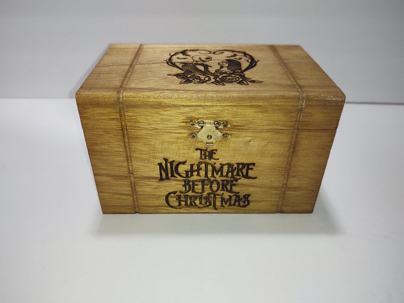 May include: A wooden box with a hinged lid and a brass latch. The box is decorated with an engraved design of Jack Skellington and Sally from the movie "The Nightmare Before Christmas". The text "The Nightmare Before Christmas" is also engraved on the front of the box.