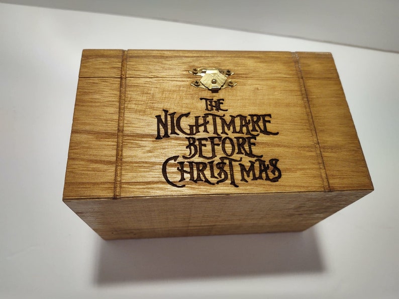 May include: A wooden box with a hinged lid and a brass latch. The box is decorated with a black burned-in design that reads "The Nightmare Before Christmas".