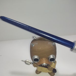 Op de afbeelding: Een blauwe lightsaber met zilveren accenten en een glinsterende afwerking. De lightsaber wordt vastgehouden door een bruin en beige figuur met grote ogen.