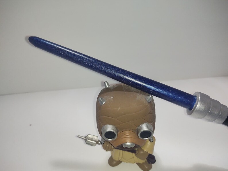 Op de afbeelding: Een blauwe lightsaber met zilveren accenten en een glinsterende afwerking. De lightsaber wordt vastgehouden door een bruin en beige figuur met grote ogen.