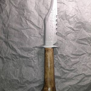 Puede incluir: Un cuchillo plateado con mango de madera marrón. La hoja tiene un borde dentado y un diseño decorativo. El mango tiene forma de hueso.