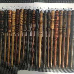 Pu&ograve; includere: Venti bacchette di legno con vari disegni e finiture, etichettate dalla A alla T. Le bacchette sono disposte in fila su una superficie bianca con un motivo a griglia nera.