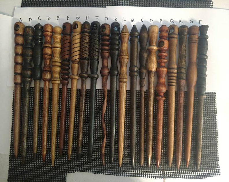 Pu&ograve; includere: Venti bacchette di legno con vari disegni e finiture, etichettate dalla A alla T. Le bacchette sono disposte in fila su una superficie bianca con un motivo a griglia nera.