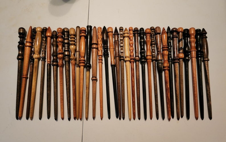 May include: A collection of 28 wooden wands, each with a unique design and finish. The wands are arranged in a row on a white surface.