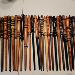 May include: A collection of 28 wooden wands, each with a unique design and finish. The wands are arranged in a row on a white surface. Some wands are dark brown, while others are light brown or have a reddish hue.