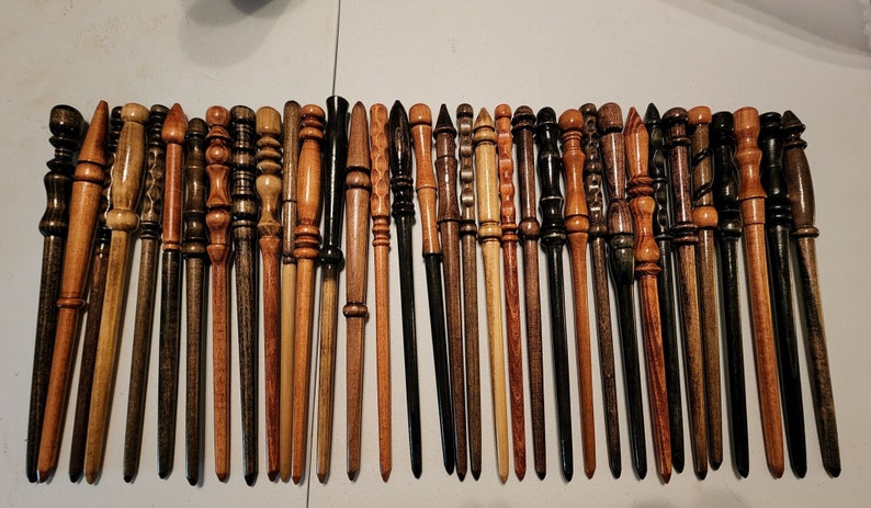 May include: A collection of 28 wooden wands, each with a unique design and finish. The wands are arranged in a row on a white surface. Some wands are dark brown, while others are light brown or have a reddish hue.