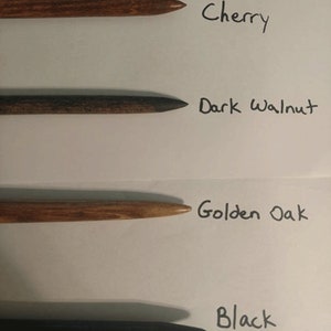 Pu&ograve; includere: Quattro pioli di legno in diverse macchie di legno: ciliegio, noce scuro, rovere dorato e nero. I pioli sono disposti verticalmente su una superficie bianca con i nomi delle macchie di legno scritti accanto a ciascun piolo.